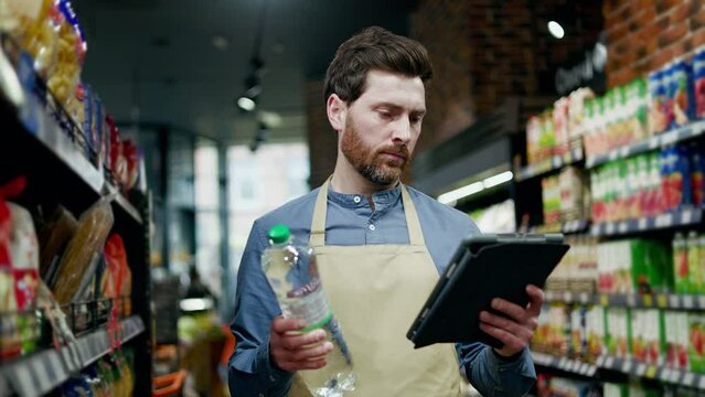 Attentive salesperson in uniform standing in food department with bottle of water and professional tablet in hands. Concerned manager inspecting products for quality and compliance with requirements.