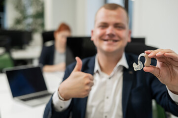 Portrait of a man holding a hearing aid and showing thumbs up in the office. 