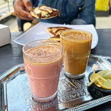 Closeup shot of Kashmiri chai tea poured in a glass cup alongside Doodh Patti Chai Milk Tea served on a silver platter with man having chocolate calzone stuffed naan in the background.