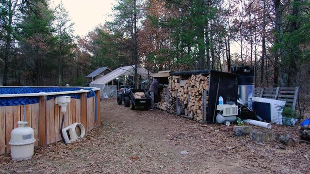 Wide angle shot of man stocking woodshed with firewood collected from processing side to be stored and used through winter days in cast iron stove to heat house.