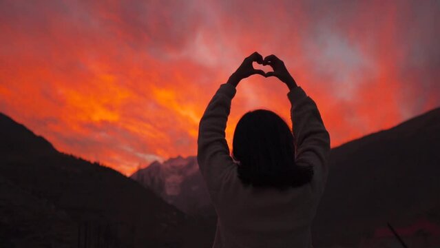 Teenager girl standing against snowy Himalaya mountains and making heart shape with fingers. Fiery red clouds during sunset at Keylong, Lahaul, India. Vacation, tourism and Summer holidays concept.