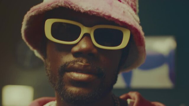 Zoom-in closeup portrait of stylish African American hipster guy posing for camera indoors wearing pink plushy bucket hat and yellow glasses