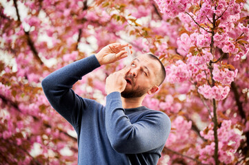 Man allergic using medical eyes drops, suffering from seasonal allergy at spring in blossoming...