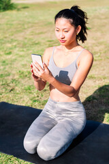 young asian woman in sportswear kneeling on her mat using mobile phone for a yoga session on the park grass, technology and healthy lifestyle concept
