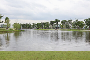 Panoramic view of public park with green grass, beautiful lake and fountain along with shrubs and trees on summer cloudy day. Image use for meteorology forecast and travel destination background.