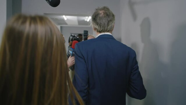 Angry politician quickly walks down the hallway of European Parliament and refuses to give interview to journalists. Busy diplomat or minister surrounded by crowd of TV news press. Hot button issues.