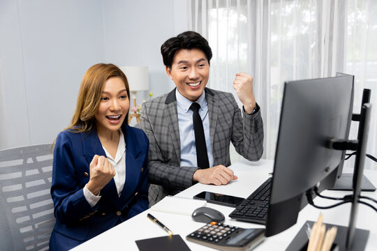 Successful smiling Asian business partners raising fist up together in profitable project job in stock exchange database on pc wearing formal suit at modern home office at side view image. Infobahn.