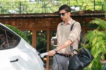 Eco friendly man and sustainable urban commute with EV electric car recharging at outdoor coffee...