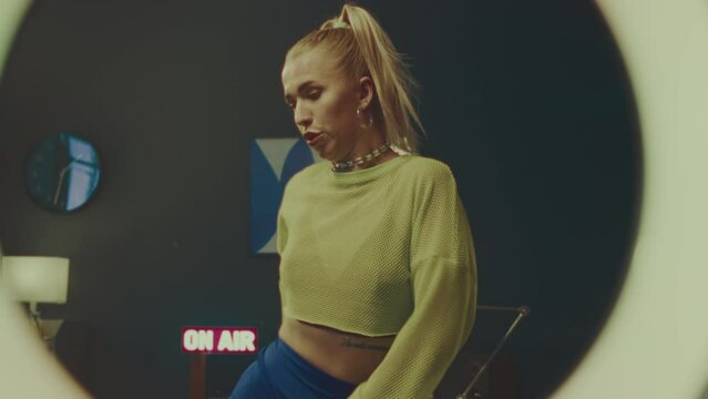 Through LED ring lamp medium slowmo of young attractive blonde girl wearing neon yellow semi sheer long sleeve crop top, chocker and hoop earring dancing hip hop indoorsThrough LED ring lamp medium sl
