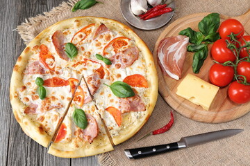On a wooden table is a traditional pizza with ham and tomatoes next to a cutting board with ingredients. 