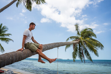 Mężczyzna na rajskiej plaży siedzi na palmie - obrazy, fototapety, plakaty