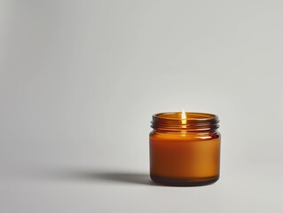 A candle is lit in a glass jar. The jar is brown and has a clear lid. The candle is the main focus of the image, and it creates a warm and cozy atmosphere