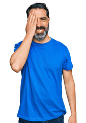 Middle aged man with beard wearing casual blue t shirt covering one eye with hand, confident smile on face and surprise emotion.