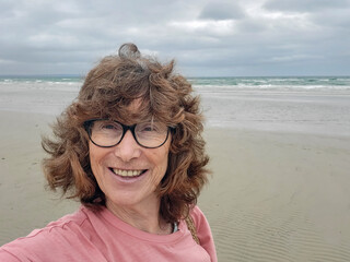 Mature woman takes a selfie while walking on Mordiallic Beach. She is smiling and wearing a casual t-shirt.