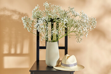 home decor and design concept - close up of gypsophila flowers in vase and straw hat on vintage chair over beige background - 767716425