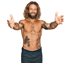 Handsome man with beard and long hair standing shirtless showing tattoos looking at the camera smiling with open arms for hug. cheerful expression embracing happiness.