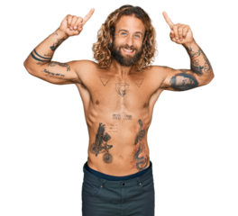 Handsome man with beard and long hair standing shirtless showing tattoos smiling amazed and surprised and pointing up with fingers and raised arms.