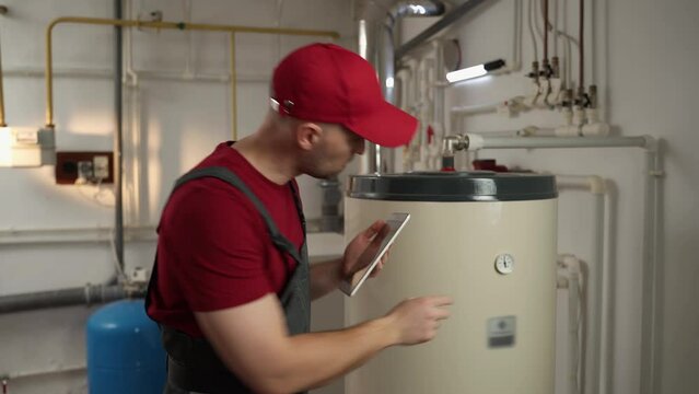 A qualified plumber in a red cap and overalls inspects a household water heater in the basement of the house, holding a tablet for notes and carefully checking the system for the need for maintenance