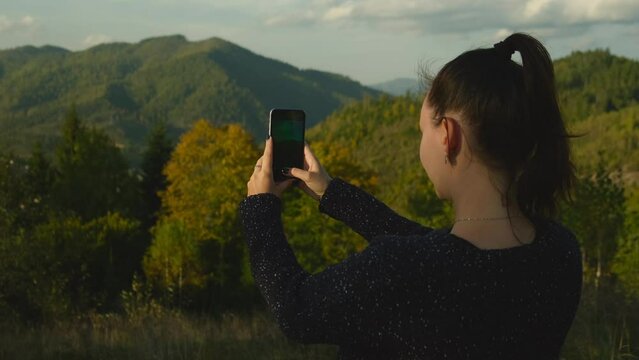 Happy girl making photos of mountains on cellphone camera. Young woman standing on top of mountain peak and photographing scenic landscape using mobile phone device
