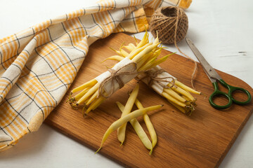 Cutting board with two bunches and several pods of flat runner bean pods heap, yellow kitchen towel, scissors and ball of thread  on white wooden background. .