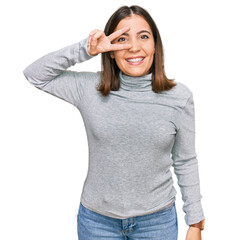 Fototapeta premium Young beautiful woman wearing casual turtleneck sweater doing peace symbol with fingers over face, smiling cheerful showing victory