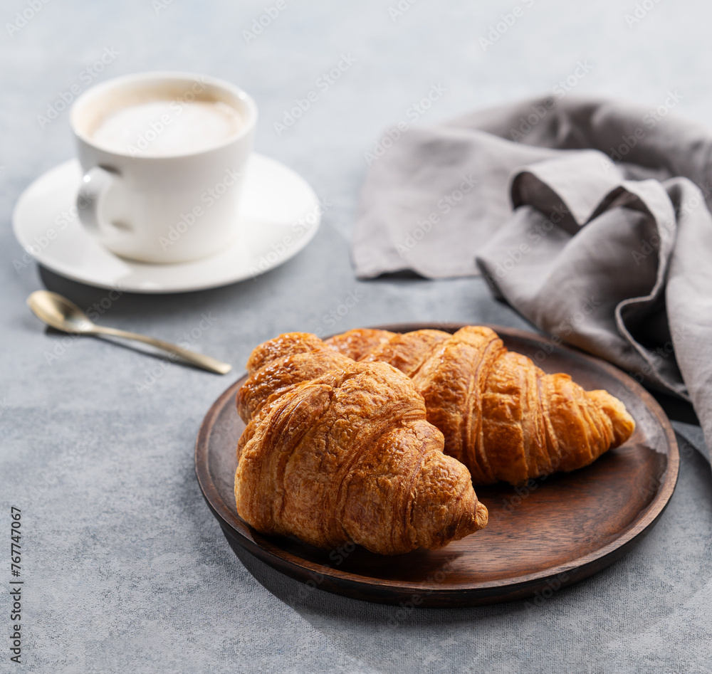 Wall mural A cup of aromatic cappuccino coffee and fresh croissants in a wooden plate on a blue background with morning light.