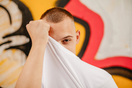 A man with a shaved head is hiding his face behind a white shirt. The man's face is angry and he is clenching his fist
