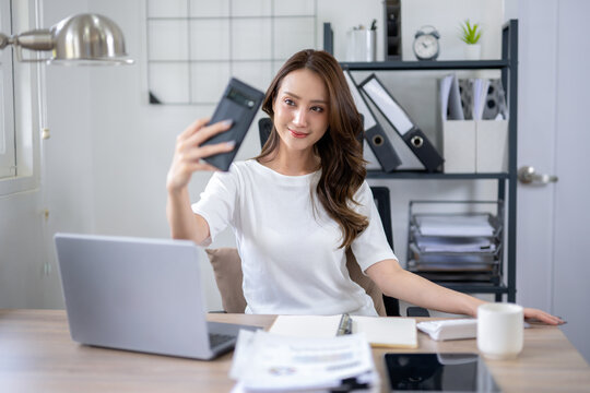 A young woman takes a break for a selfie, in a home office setup with a laptop and documents.