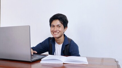 Young handsome Asian male student is happy studying at a table using a book and laptop