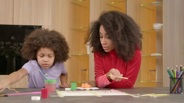 Happy inspired cute school age African American daughter choosing paintbrush while enjoying leisure together with pretty black mother, painting picture on paper with colorful paints indoors.