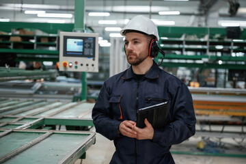 Machine behind, holding notepad. Factory worker is indoors with hard hat