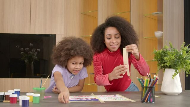 Cheerful adorable elementary age black girl and happy affectionate pretty African American mother finishing creating picture on paper , detaching ready painting from table at home.
