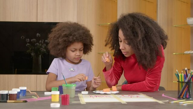 Caring loving attractive black mother teaching lovely elementary age curly African American daughter to sponge painting techniques, developing creativity and artistic skills of happy child indoors.