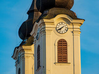 Glockenfenster, Turmuhren und Holzdächer der zwei Kirchtürme des Benediktinerklosters in Tihany am Plattensee in Ungarn im warmen Licht der tief stehenden Sonne - 767787494