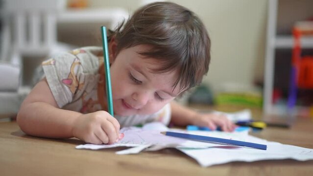 child draws with pencils on a sheet. happy family child dream concept. baby girl lies on the floor indoors and draws pictures , developing fine motor skills. lifestyle drawing of a child