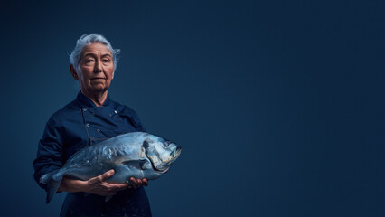 An elderly chef in a blue uniform holds a sizable fish against a dark blue backdrop, showcasing a culinary theme