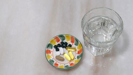 A small plate with various health supplement pills and capsules. On a marble surface, with a glass of water beside with plate.
