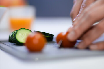 Obraz na płótnie Canvas Close up shot of female hands preparing breakfast in kitchen using knife. Woman slicing fresh cucumbers and tomatoes on cutting board making a salad indoor. Food concept