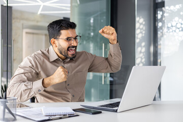 Successful businessman inside office celebrating victory and triumph, man reading happy news from laptop, entrepreneur working at workplace, satisfied with achievement results - obrazy, fototapety, plakaty