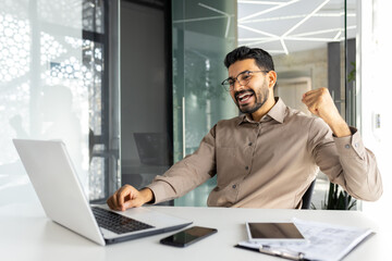 Fototapeta na wymiar Successful businessman inside office celebrating victory and triumph, man reading happy news from laptop, entrepreneur working at workplace, satisfied with achievement results