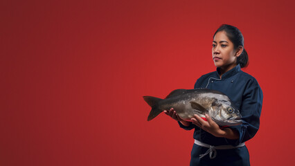 A middle-aged chef in a dark navy uniform gently holds a large fish while posing against a plain red backdrop
