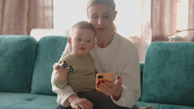 Mum sitting with her son kid child infant toddler boy and using smartphone phone for shopping online, typing messages, browsing internet, scrolling social media