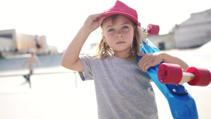 child with a skateboard. girl in a red cap with a skateboard on the playground portrait. skateboarder child close-up outdoors sun glare lifestyle. kid skateboarder looking at the camera - 767831282