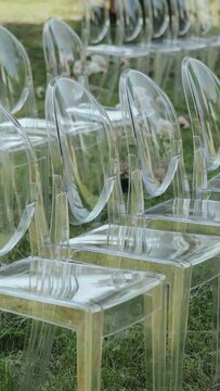 Clear plastic chairs arranged on vibrant grass field prepared for wedding ceremony in the park.VERTICAL VIDEO. Chairs on green lawn, slow motion.