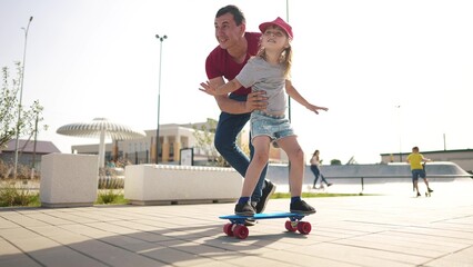 learn to skateboard. dad teaches daughter to ride a skateboard outdoors at the playground. father and daughter play training concept. parent teaching child daughter to lifestyle skateboard