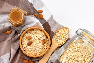 Oatmeal in a wooden bowl with almonds and pieces of banana ready to cook, honey, scattered oat flakes and spoon on white background. View from above. - 767840224