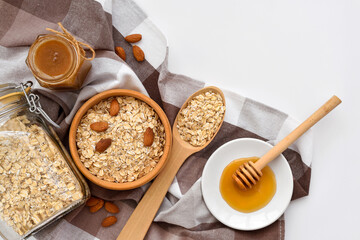 Oatmeal in a wooden bowl with almonds and pieces of banana ready to cook, honey, scattered oat flakes and spoon on white background. View from above. Place for text - 767840225