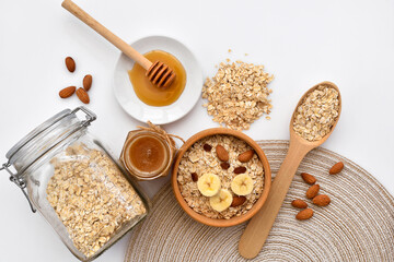 Oatmeal in a wooden bowl with almonds and pieces of banana ready to cook, honey, scattered oat flakes and spoon on white background. View from above. Place for text - 767840255