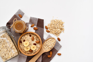 Oatmeal in a wooden bowl with almonds and pieces of banana ready to cook, honey, scattered oat flakes and spoon on white background. View from above. Place for text - 767840287