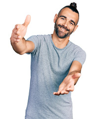 Hispanic man with ponytail wearing casual grey t shirt looking at the camera smiling with open arms...